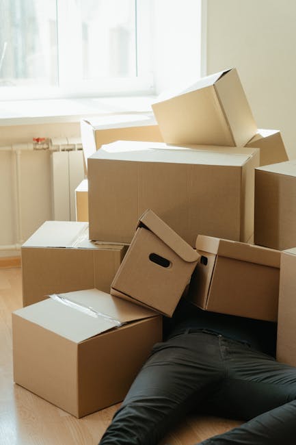 A young woman sitting cross-legged on a wooden floor inside a residential property, surrounded by numerous large cardboard moving boxes of various sizes with printed labels and red packing tape. She i