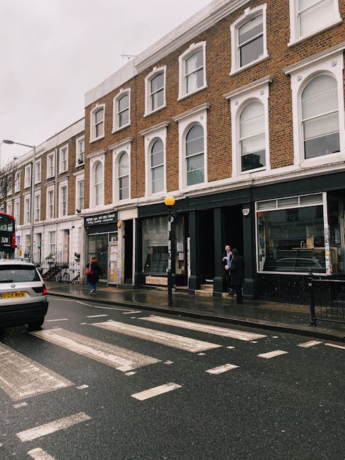 The image shows the exterior of a three-storey brick building on Gloucester Road in South Kensington, with large ground-floor windows and white decorative window surrounds on the upper floors. The pavement in front is wet, suggesting recent rain, and features a black lamppost with a yellow globe light. Several pedestrians, some wearing face masks, are walking along the sidewalk, while a white van and a red double-decker bus are visible on the rainy street. A white vehicle with a UK license plate is parked on the road, and a street zebra crossing is in the foreground. This scene captures a typical busy day during a home relocation or moving service, with South Kensington Man and Van likely involved in logistics and transportation for house removals, as suggested by the context and setting.