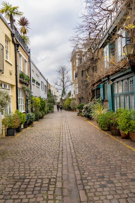 A narrow cobblestone street in South Kensington lined with white terraced houses featuring black wrought iron balconies and window boxes filled with plants. Several potted trees and shrubs are placed