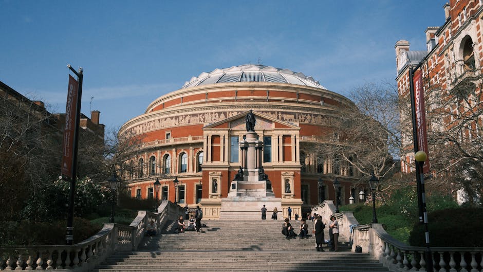 Inside the grand interior of the Royal Albert Hall, a large suspended whale skeleton with a detailed skull and ribcage hangs from the high glass ceiling supported by steel beams. The hall's walls are 