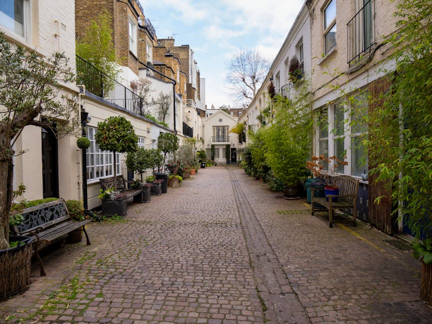 A narrow cobblestone street within a residential area in South Kensington, lined with white-painted terraced houses that feature large windows and black wrought-iron balconies. The buildings are decor